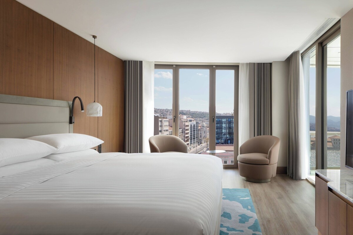 A hotel room with a large bed, two armchairs, a TV, and a city view through large windows.