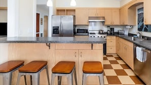 A modern kitchen with wooden cabinets, a granite countertop, stainless steel appliances, and a checkered floor. - Raven Lodge, Amazing Location and Epic Views, Sleeps 10 (Seward)