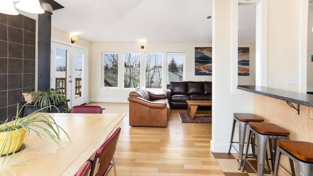 A modern kitchen with a wooden countertop and bar stools, a seating area with a sofa and a coffee table, and large windows offering a view of trees and mountains. - Raven Lodge, Amazing Location and Epic Views, Sleeps 10 (Seward)