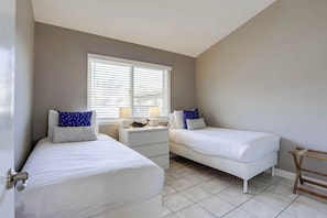 A bedroom with two single beds, a nightstand, a window with blinds, and a wooden rack.