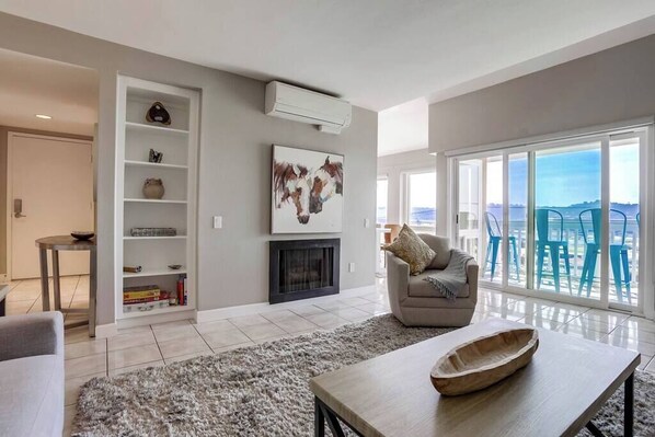 A modern living room with a fireplace, a bookshelf, a sofa, and a view of the sea through large windows.