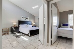 Um quarto com uma cama grande, uma cama menor, uma mesinha de cabeceira, uma luminária, um mapa-múndi na parede e uma porta que dá para outro cômodo.