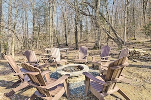 A circular seating arrangement with Adirondack chairs around a central fire pit, set in a forested area with trees and logs in the background.