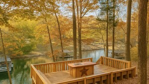 Eine Holzterrasse mit einem sternförmigen Tisch, mit Blick auf einen ruhigen Fluss, der von Herbstbäumen gesäumt wird.