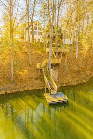 A house with a wooden dock extending into a calm body of water, surrounded by trees and autumn leaves.