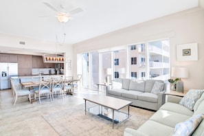 A spacious living room with a large window, a dining area, and a kitchen in the background.