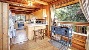 L’intérieur d’un chalet en bois comprend une cuisine, des tabourets de bar et une grande fenêtre donnant sur un paysage verdoyant.