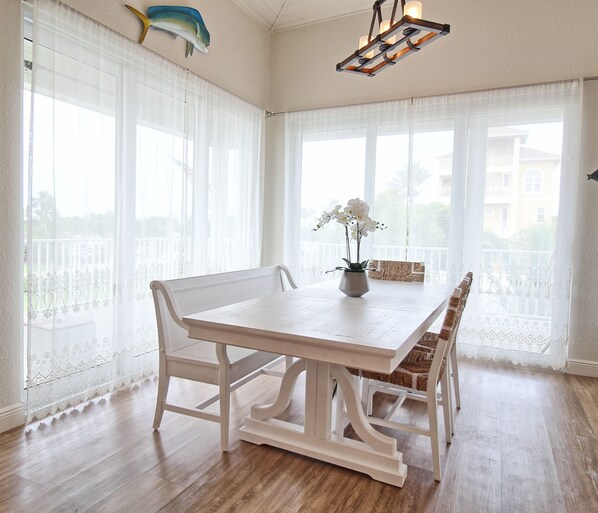 A dining area with a white table and chairs, a vase with flowers, and large windows with sheer curtains.