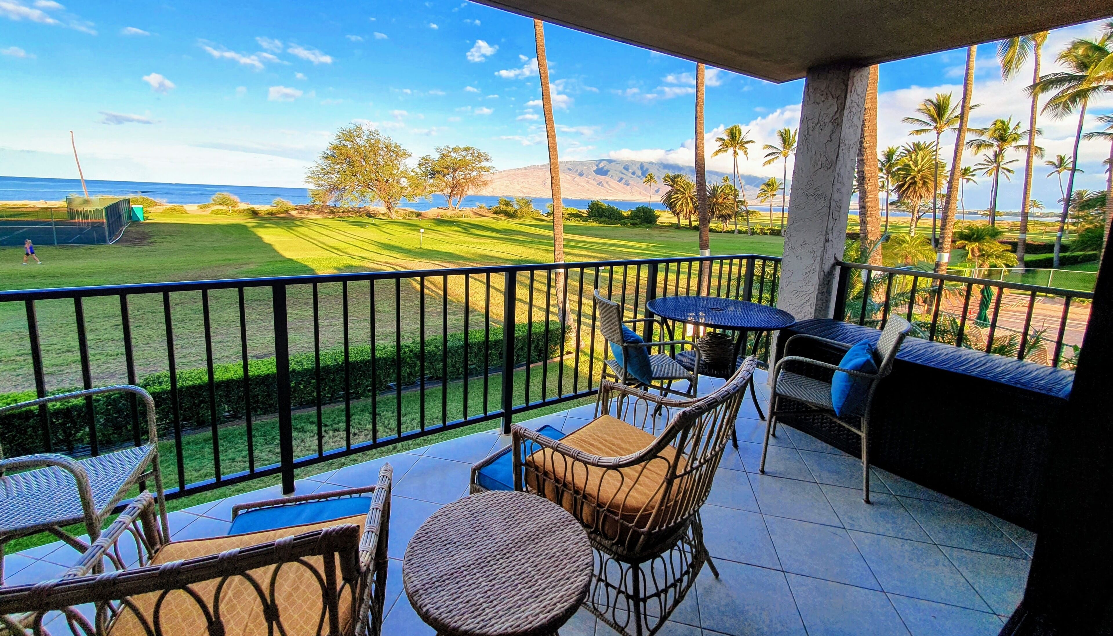 A balcony with wicker furniture, a view of a golf course, and palm trees.