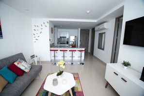 Uma sala de estar moderna com um sofá cinzento, almofadas coloridas, uma mesa de centro branca com um vaso de flores e um tapete vibrante.