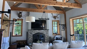 Un salón amplio con techo alto, vigas de madera a la vista, chimenea y ventanas grandes con vistas al exterior.