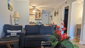 A living room with a dark grey sofa, a small table with a plant, and a coffee table with books. There is a kitchen in the background with cabinets and a microwave. - The Zen Place<br>Weekly and Monthly discount (Hilton Head Island)