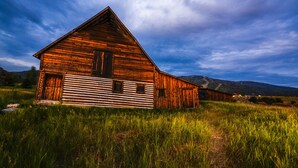 A wooden barn with a sloped roof, surrounded by tall grass and mountains in the background.