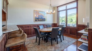 A dining area with a wooden table and chairs, a chandelier, and a large window with a view of greenery.