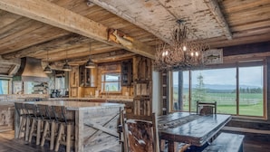 Un intérieur rustique avec une grande table à manger en bois, des tabourets de bar et une vue sur l’extérieur.