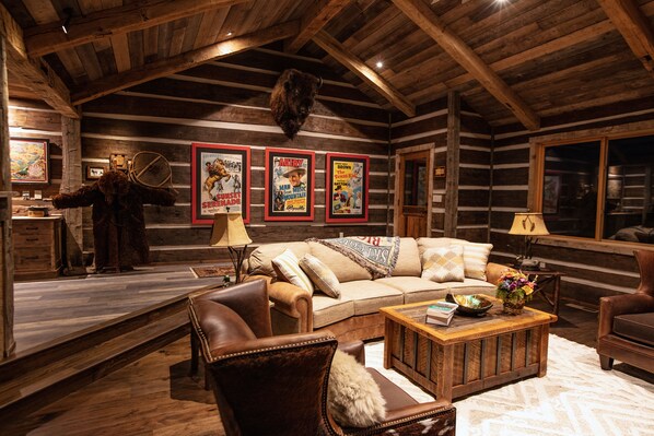 Interior de una cabaña rústica con techo de madera, un sofá, un sillón de cuero, una mesa de centro y carteles de películas enmarcados en la pared.