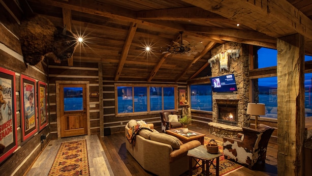 L’intérieur chaleureux d’un chalet en bois, avec une cheminée en pierre, des meubles en cuir et de grandes fenêtres offrant une vue sur le ciel nocturne.