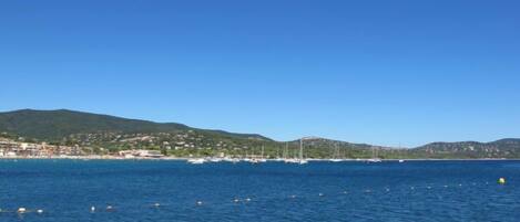 A coastal view with clear blue water, boats, and a hilly landscape in the background.