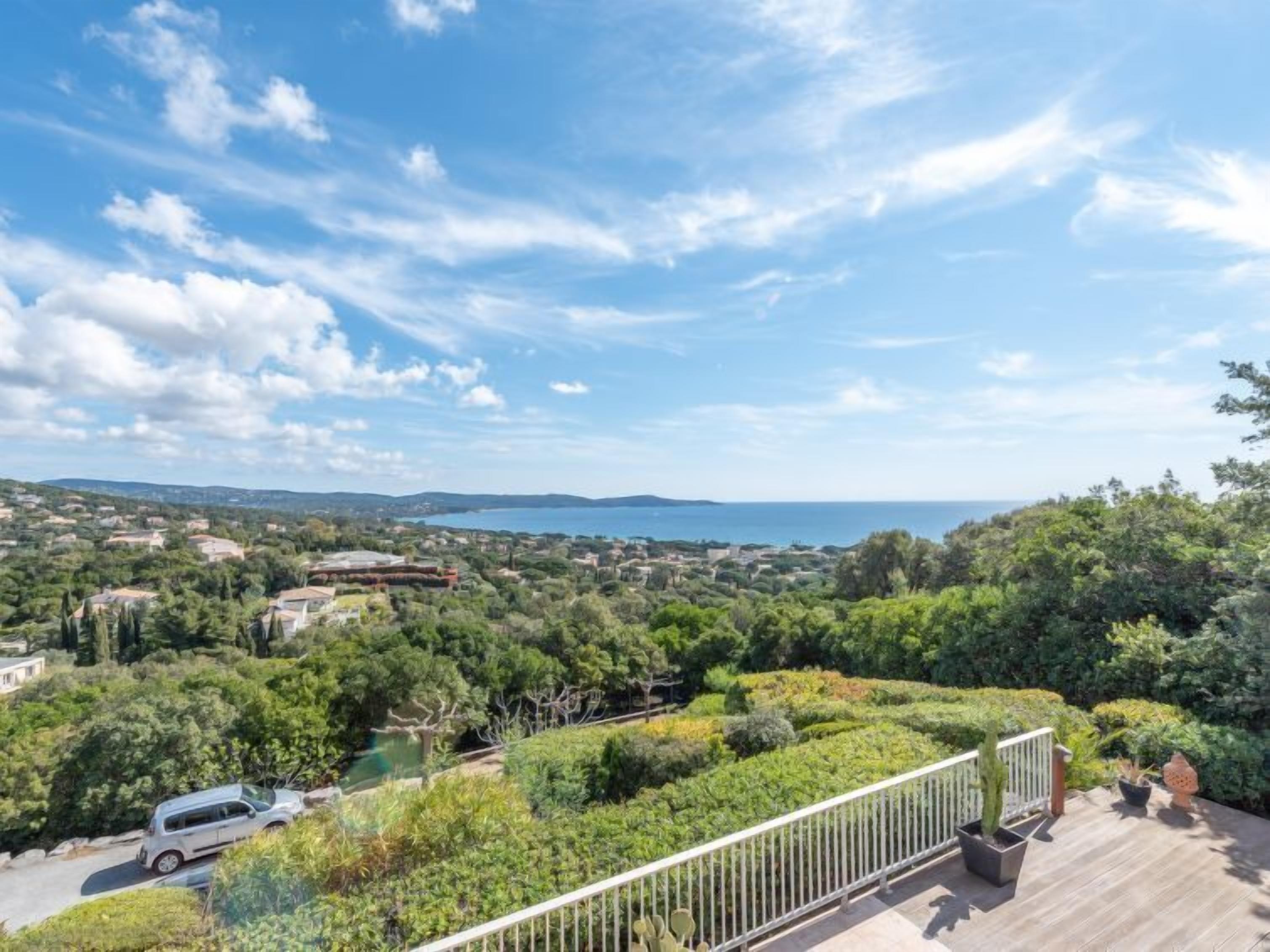 A balcony with a view of a coastal town, a car parked nearby, and a clear sky.