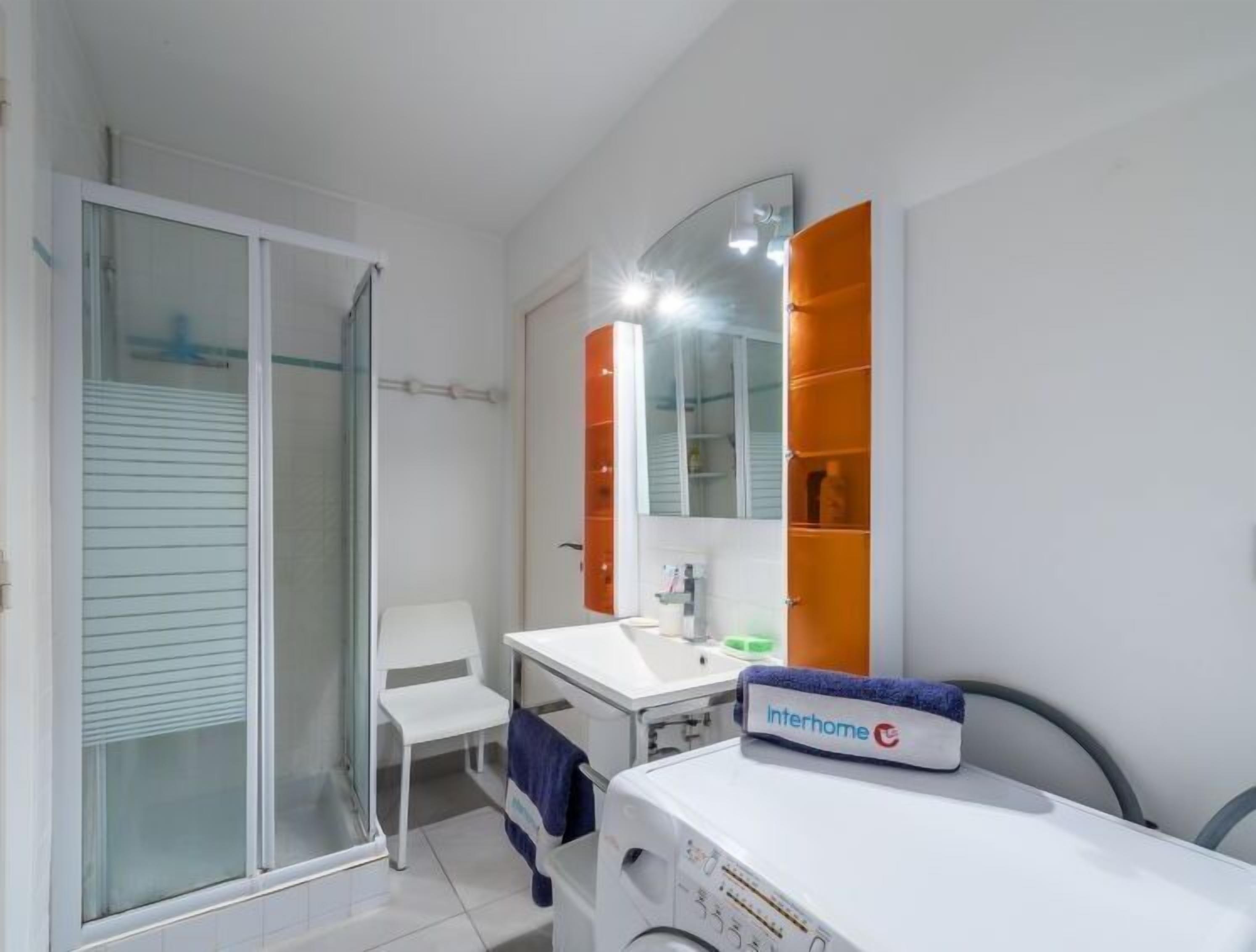 Une salle de bain moderne avec une douche à l’italienne en verre, un lavabo blanc et un grand miroir surmonté d’un meuble orange.