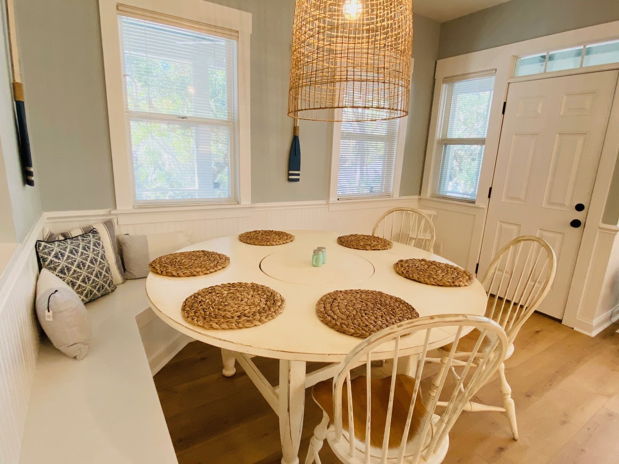 A dining area with a round table, six woven placemats, white chairs, a hanging light fixture, and a white door.