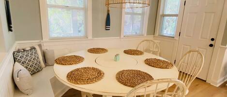 A dining area with a round table, six woven placemats, white chairs, a hanging light fixture, and a white door.