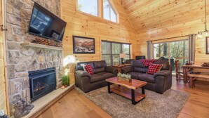 Ein gemütliches Blockhaus-Interieur mit Kamin aus Stein, zwei Ledersofas, einem Holztisch und großen Fenstern, die viel Tageslicht hereinlassen.