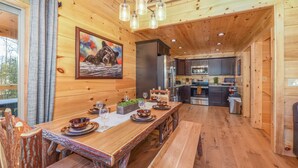 Ein Holzblockhaus-Interieur mit einem Esstisch, gedeckt für vier Personen, einer Küche mit modernen Geräten und einem großen Fenster mit Vorhängen.