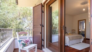 Uma varanda com uma cadeira branca e uma cesta de vime, uma porta de madeira que dá para um quarto com cama e uma pequena mesa, e uma vista de árvores e um edifício ao fundo.