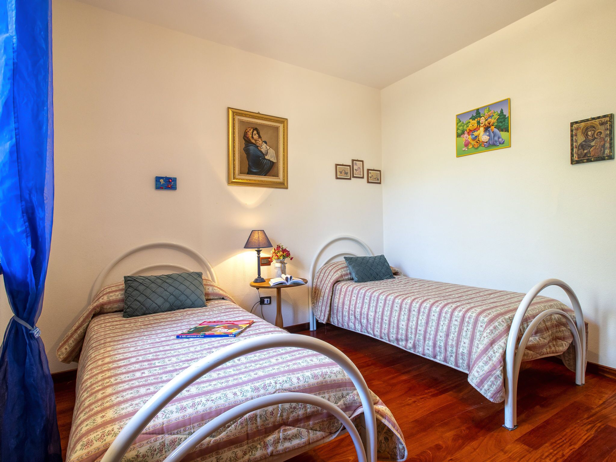 Une chambre avec deux lits simples, une table de chevet avec une lampe et une petite table sur laquelle se trouvent un livre et des fleurs.
