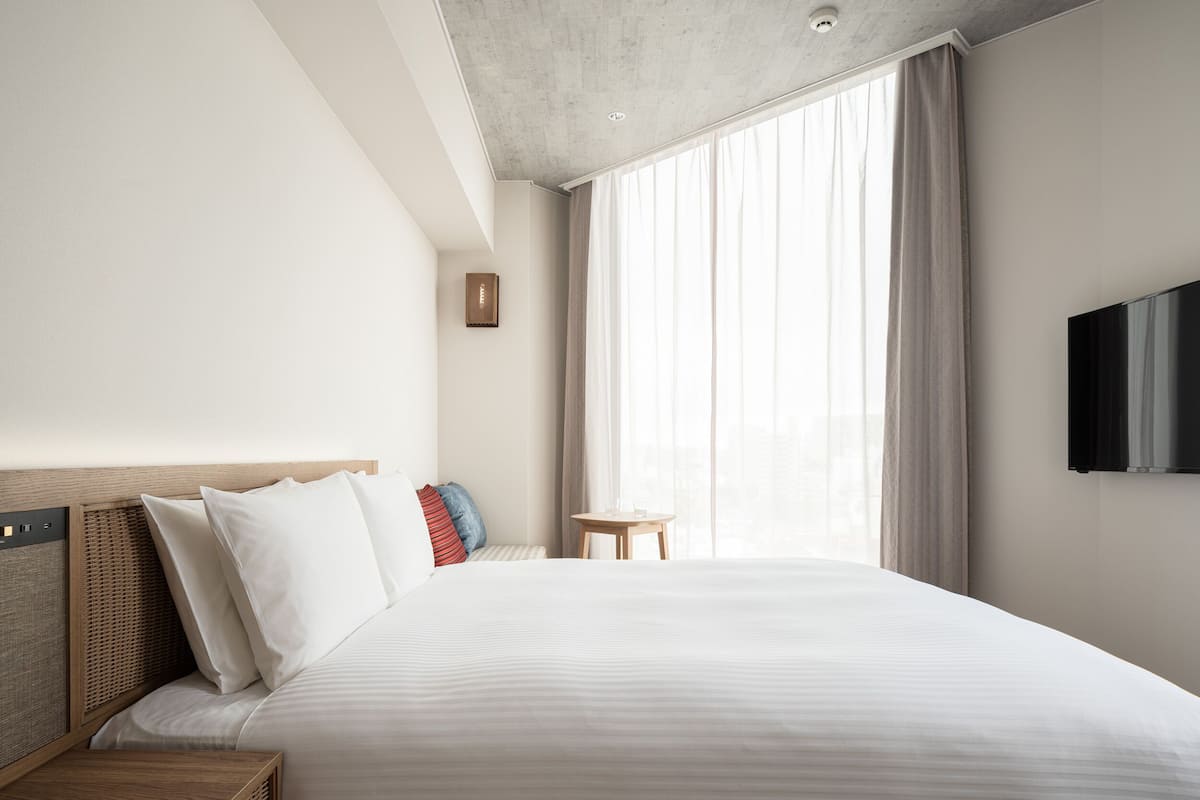 A modern hotel room with a large bed, a flat-screen TV mounted on the wall, a small round table, and a large window with sheer curtains.