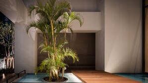 A modern indoor pool area with a wooden deck, surrounded by a glass enclosure, and a large palm tree in the center. - Nest Hotel Naha Kumoji (Naha)