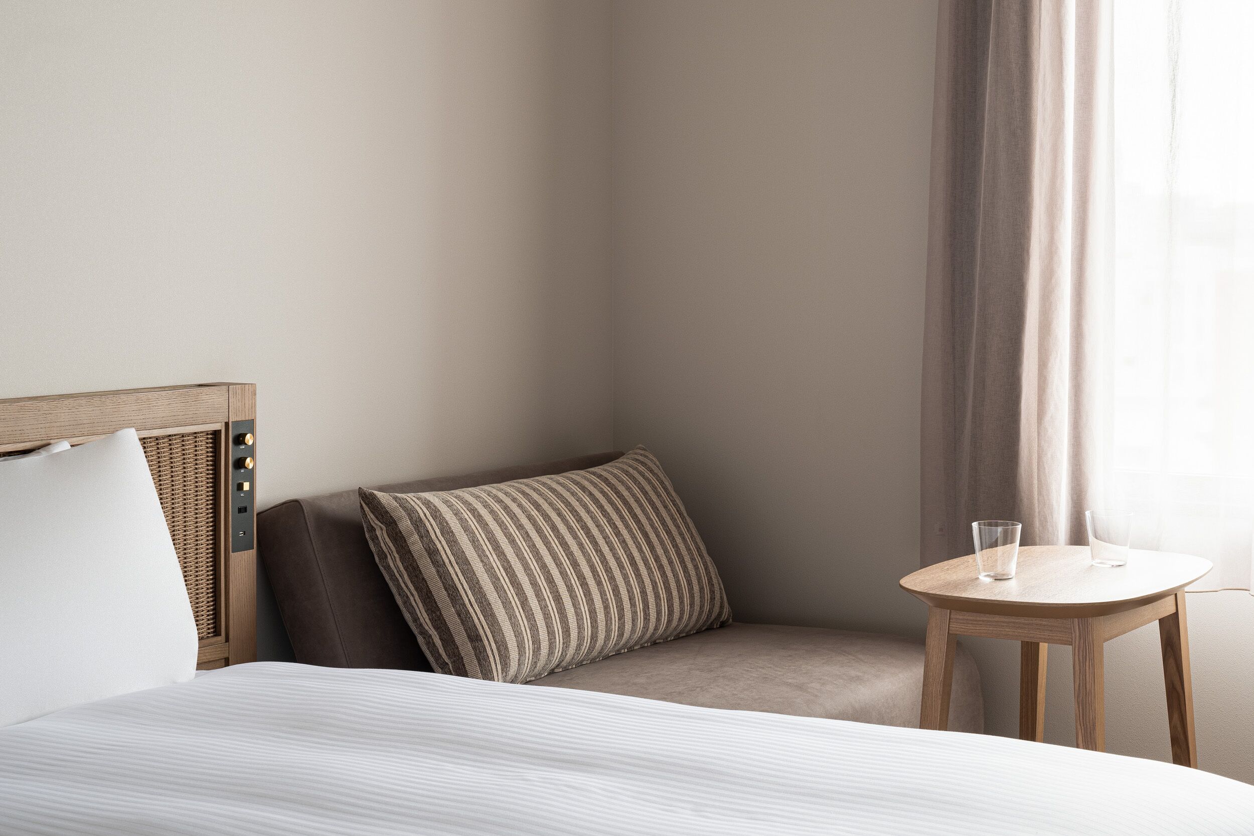 A neatly arranged hotel room with a bed, a bedside table with two glasses, a pillow, and a window with curtains.