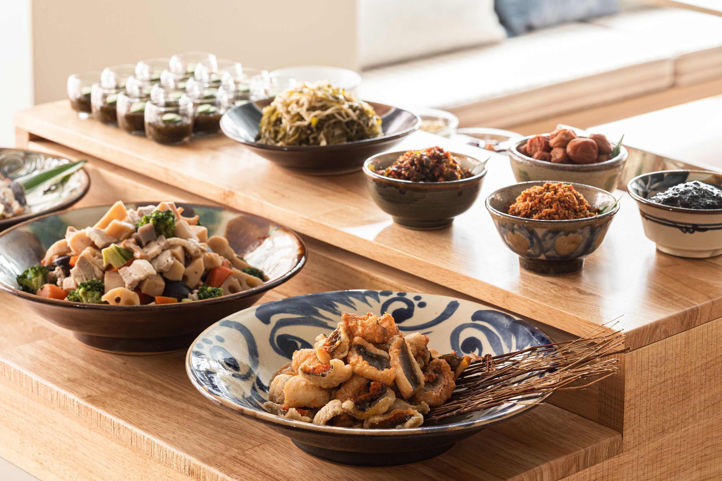 A wooden table with various dishes, including a bowl of fried food, a bowl of vegetables, and several small containers of condiments.