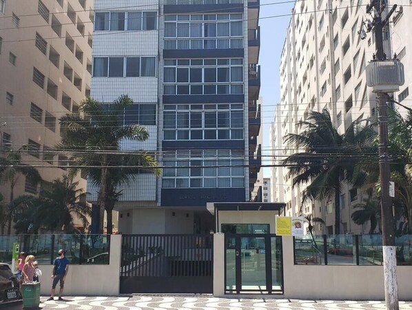 Un edificio moderno con fachada de vidrio, entrada protegida por un portón y palmeras al frente.