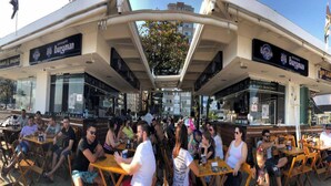 An outdoor seating area with tables and chairs, a covered section with a glass roof, and storefronts with signage. - Ed. Fte. Gonzaga beach 14and sea view (Santos)