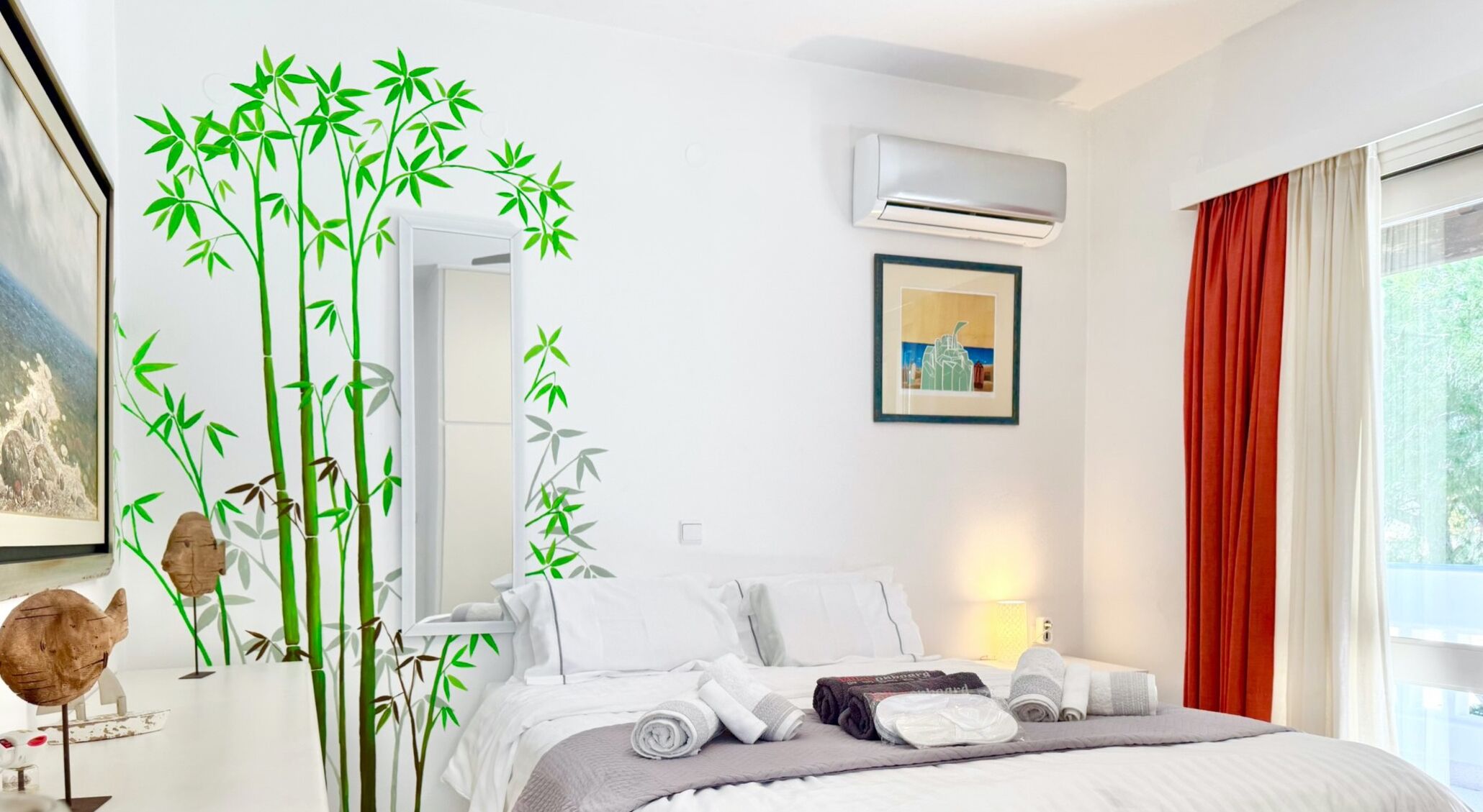 Un dormitorio con cama, almohadas, una mesita de noche, un equipo de aire acondicionado, un cuadro enmarco y una ventana con cortinas.