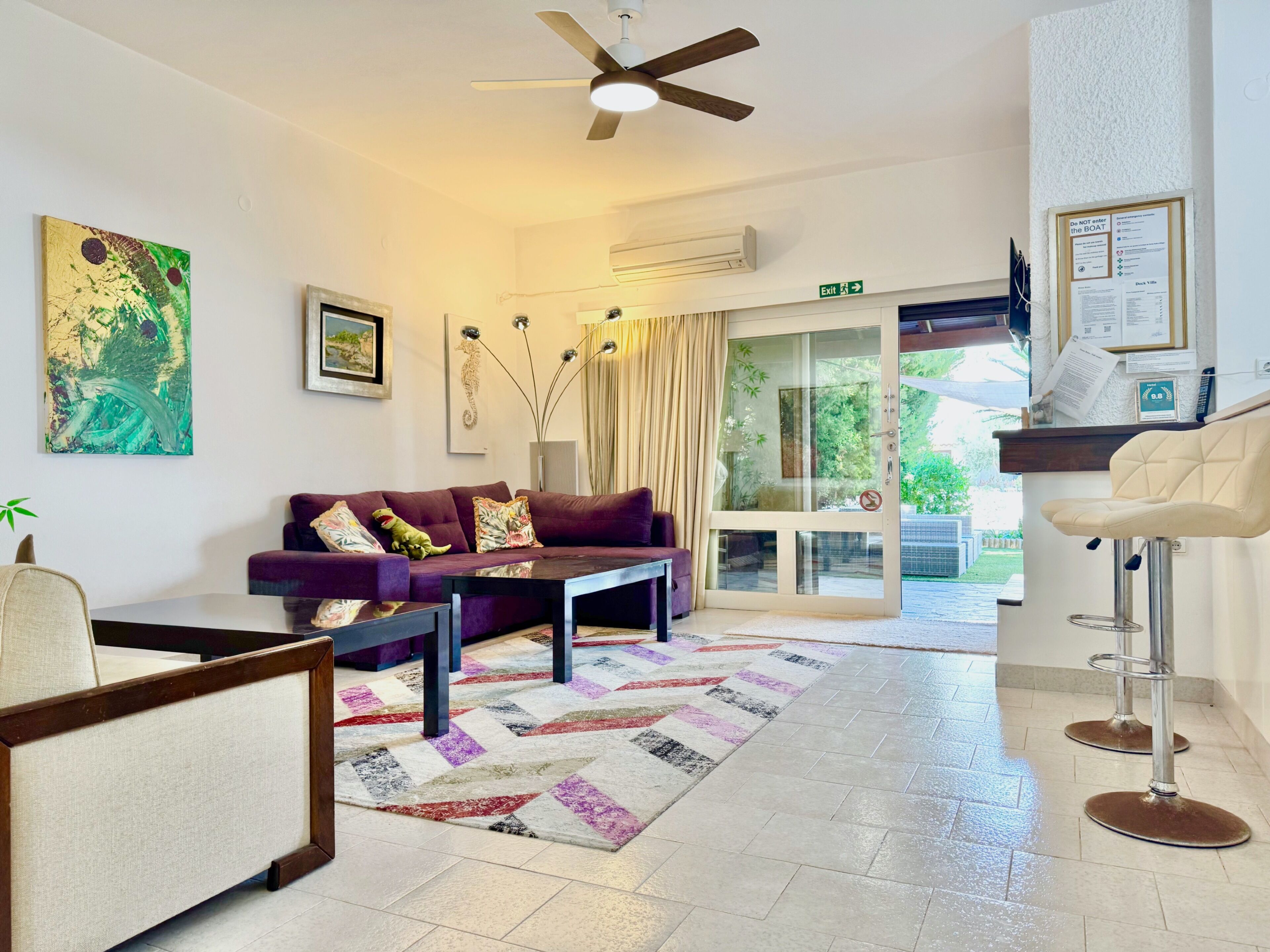 A modern hotel lobby with a purple sofa, a coffee table, a patterned rug, a ceiling fan, and a bar area with stools.