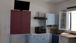 A compact kitchen with a microwave, a small oven, and a sink. There is a window with curtains and a television mounted on the wall.