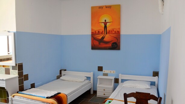 A room with two beds, a table with a chair, a wall-mounted painting, and a tiled floor.
