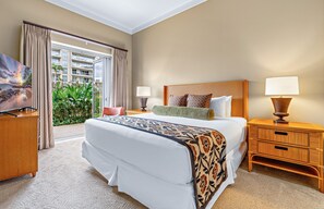 Een hotelkamer met een groot bed, twee nachtkastjes met lampen, een televisie en uitzicht op een balkon met planten.