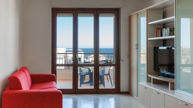A room with a red sofa, a TV on a cabinet, and a balcony with a view of the sea.