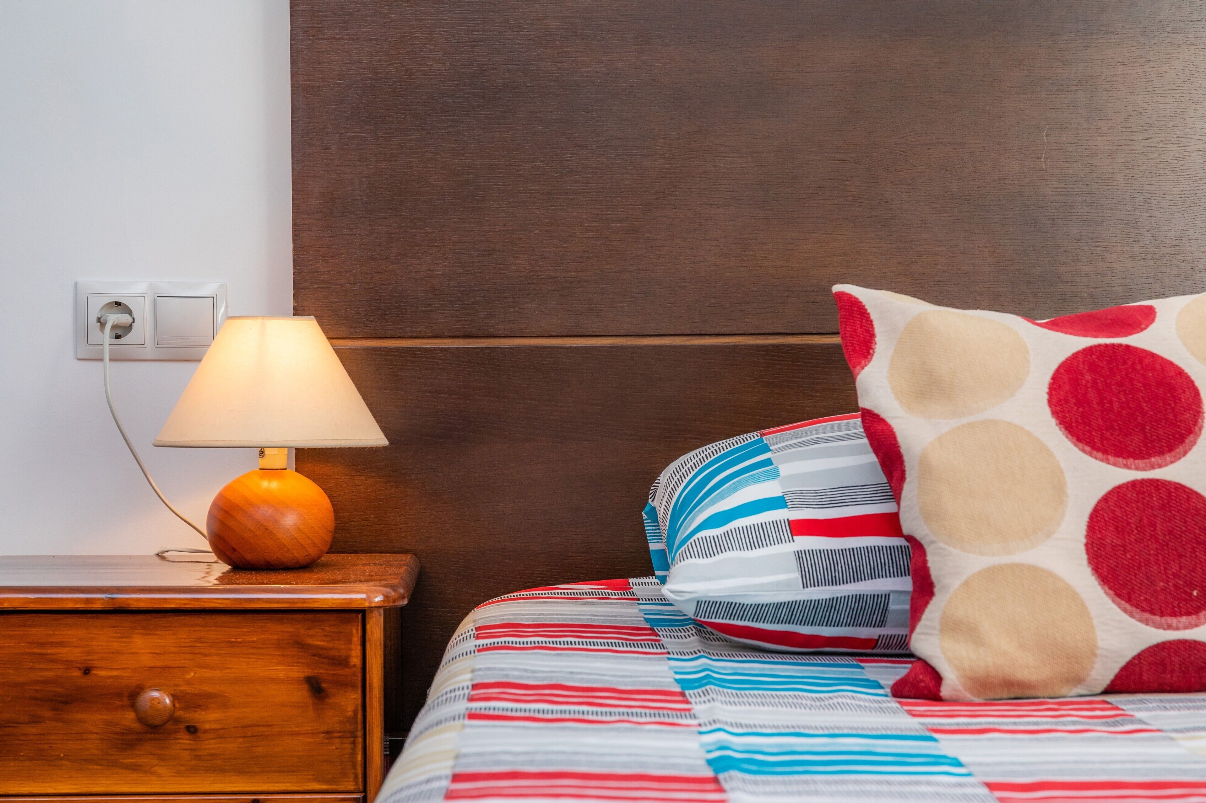 A bed with a striped and polka-dotted pillow, a wooden bedside table with a lamp, and a wall socket.