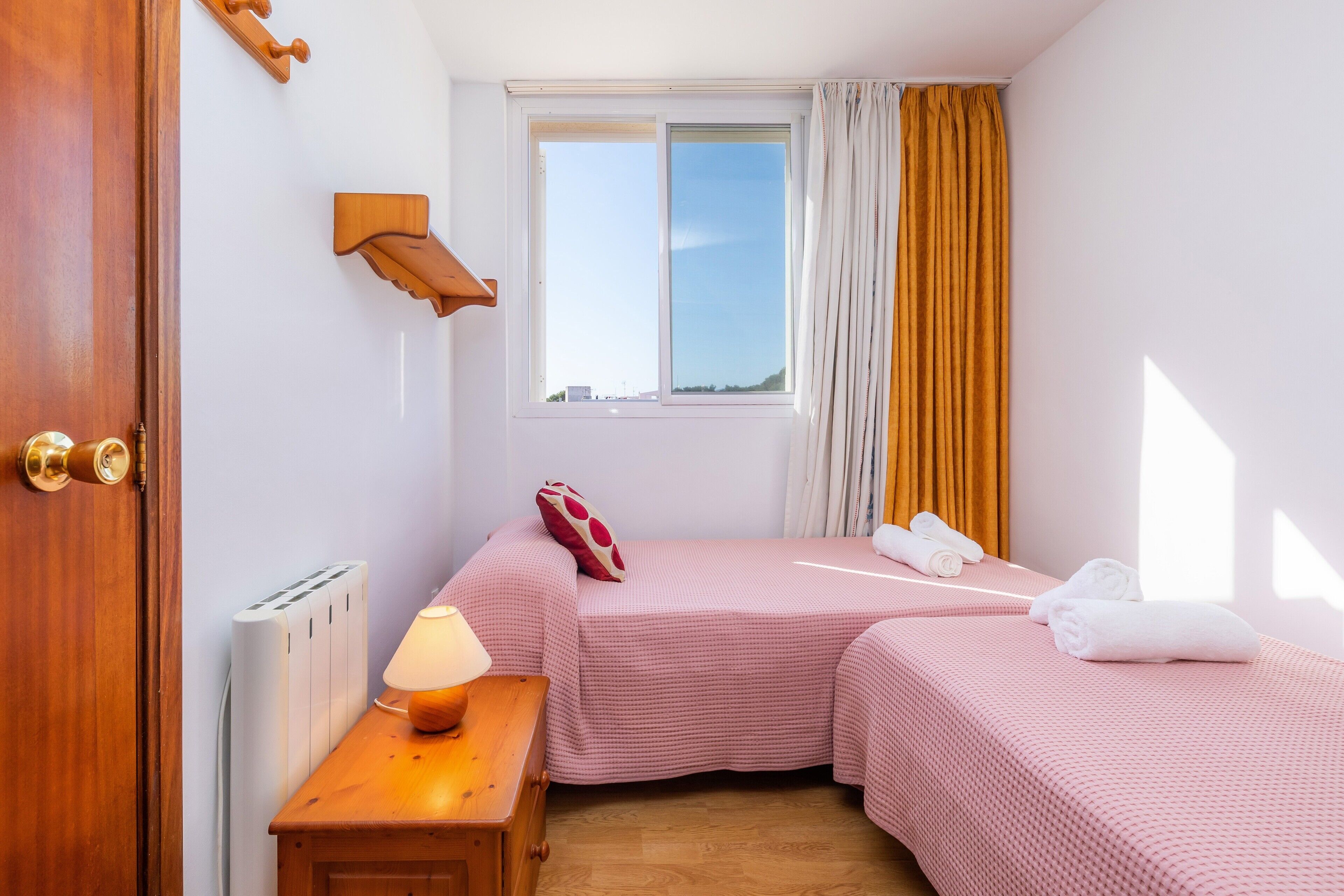 Una habitación con dos camas, una ventana, un radiador, una mesita de noche de madera y una lámpara.
