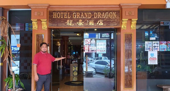 The entrance to Hotel Grand Dragon with a sign in both English and another language, a reception desk, and a glass door reflecting the street outside. - GD Hotel - Permas Jaya (Masai)