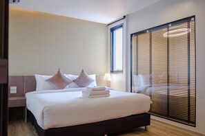 A neatly made bed with pillows and folded towels, a bedside table with a lamp, and a large window with blinds. - The Rise Suites Hotel Chiang Mai  (Chiang Mai)