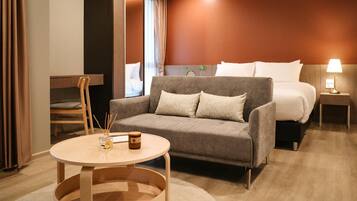 A modern hotel room with a grey sofa, a round wooden table, a bed with white bedding, and a nightstand with a lamp.