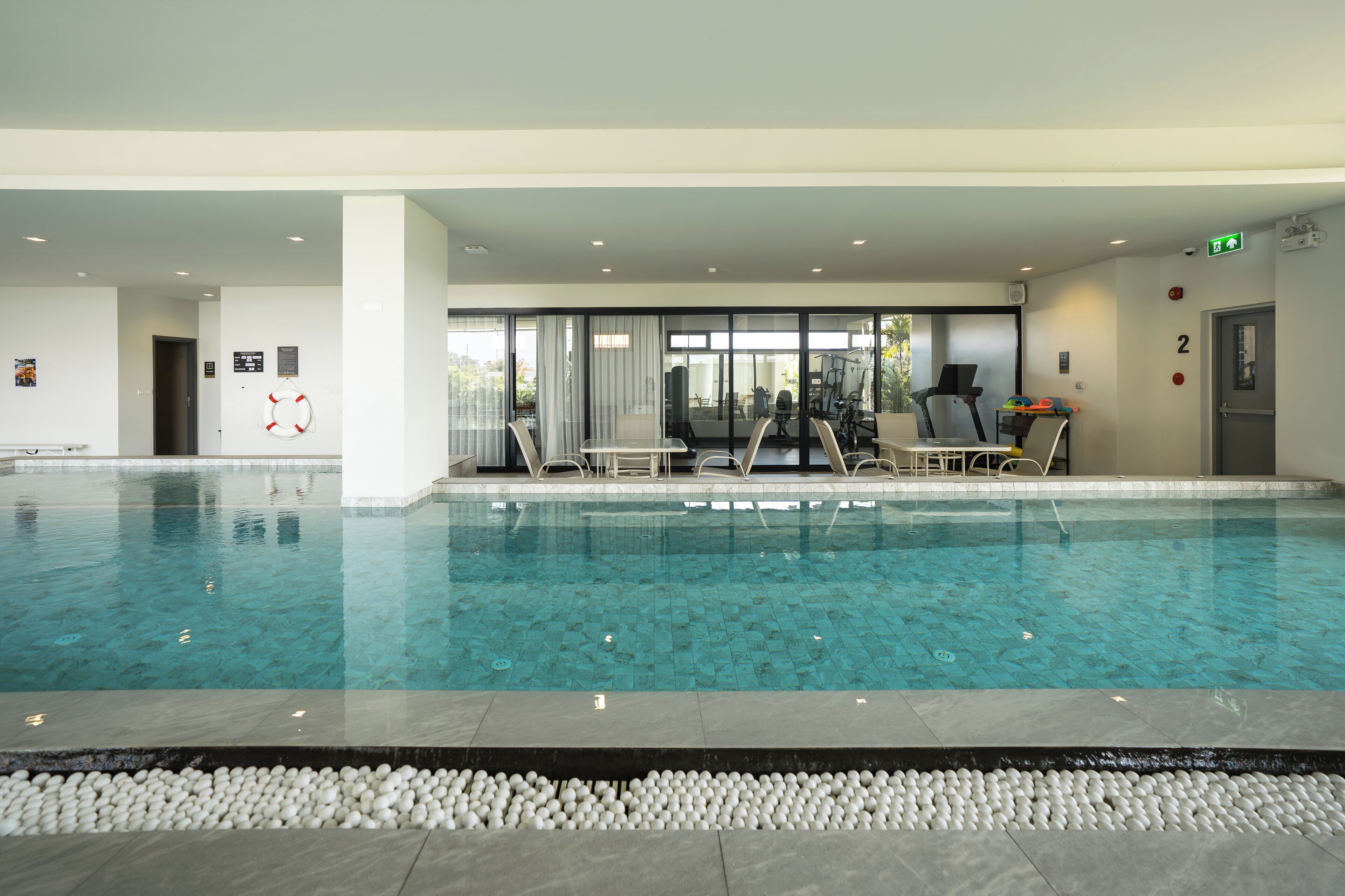 An indoor swimming pool with clear water, surrounded by a tiled deck and a seating area with chairs and tables.
