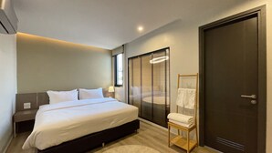 A modern bedroom with a bed, bedside table, lamp, window with blinds, and a door. - The Rise Suites Hotel Chiang Mai  (Chiang Mai)