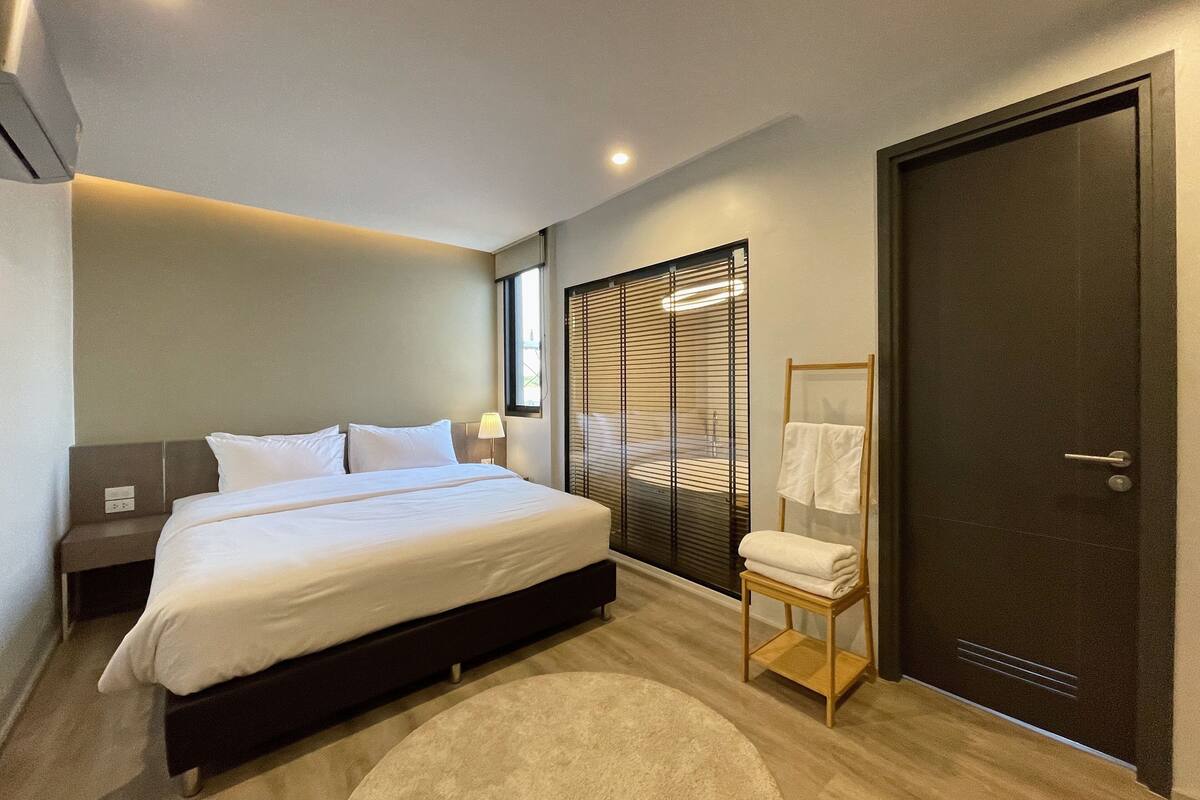 Une chambre moderne avec un lit, une table de chevet, une lampe, une fenêtre avec des stores et une porte.